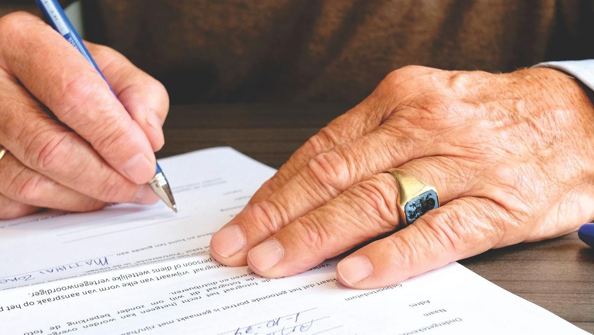 Mains âgées écrivant sur un document avec un stylo bleu sur une table en bois Mains âgées écrivant sur un document avec un stylo bleu sur une table en bois